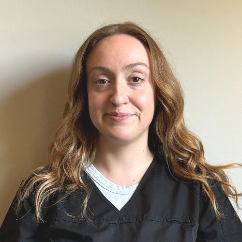 Alex A woman with wavy light brown hair, wearing a black scrub top over a gray shirt, stands against a plain light-colored wall.