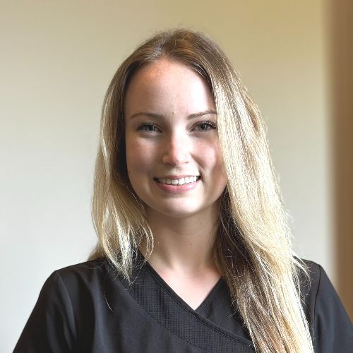 Bethany A young woman with long blonde hair, wearing a black top, is standing indoors and smiling at the camera. The background is softly lit and out of focus.