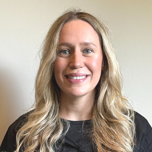 Brenna A woman with long blonde hair and blue eyes smiles at the camera. She is wearing a black top and is posed in front of a plain, light-colored background.