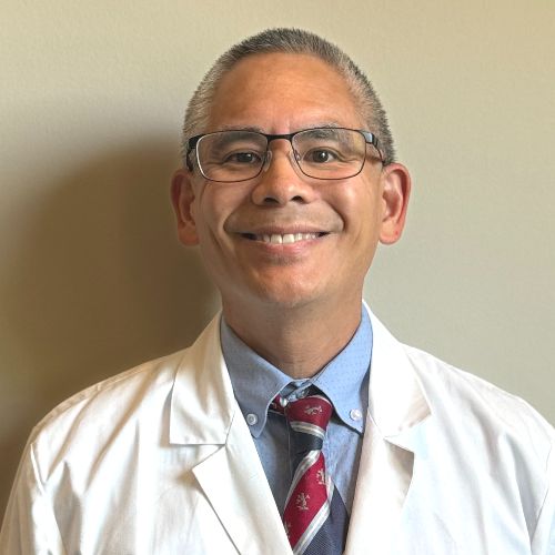 Dr Chai A man wearing glasses, a white lab coat, and a striped tie stands in front of a plain light-colored wall, smiling at the camera, ready to assist as part of the dental team.