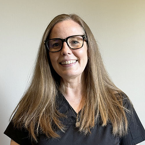 Jennifer Woman with long straight brown hair and black-framed glasses, smiling, wearing a black top, standing in front of a plain light background.