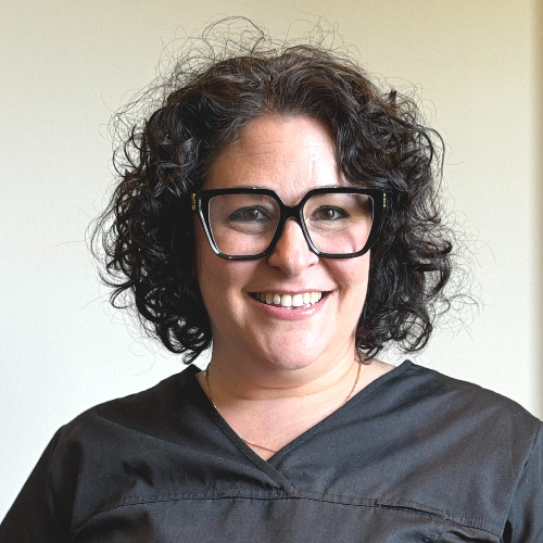 Jessica A woman with curly dark hair, large black glasses, and a black top smiles at the camera against a plain light background.