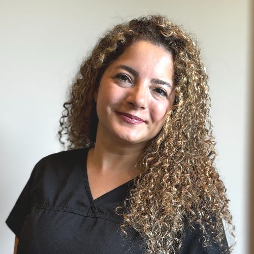Niloo A woman with long, curly hair wearing a black V-neck top stands in front of a plain, light-colored background, smiling slightly at the camera.