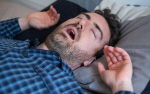 A man experiencing jaw pain in the morning while yawning in bed with his hands up.