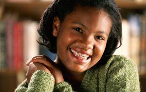 A young girl has fun with braces while smiling in front of a bookshelf.