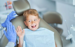 Young boy sitting in a dental chair, smiling and waving at the dentist wearing blue gloves in a clinic setting.